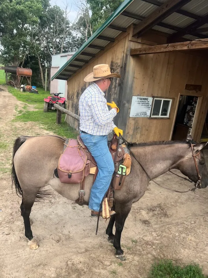 Me on a dun horse by a wooden barn, in work gloves and a straw hat.
