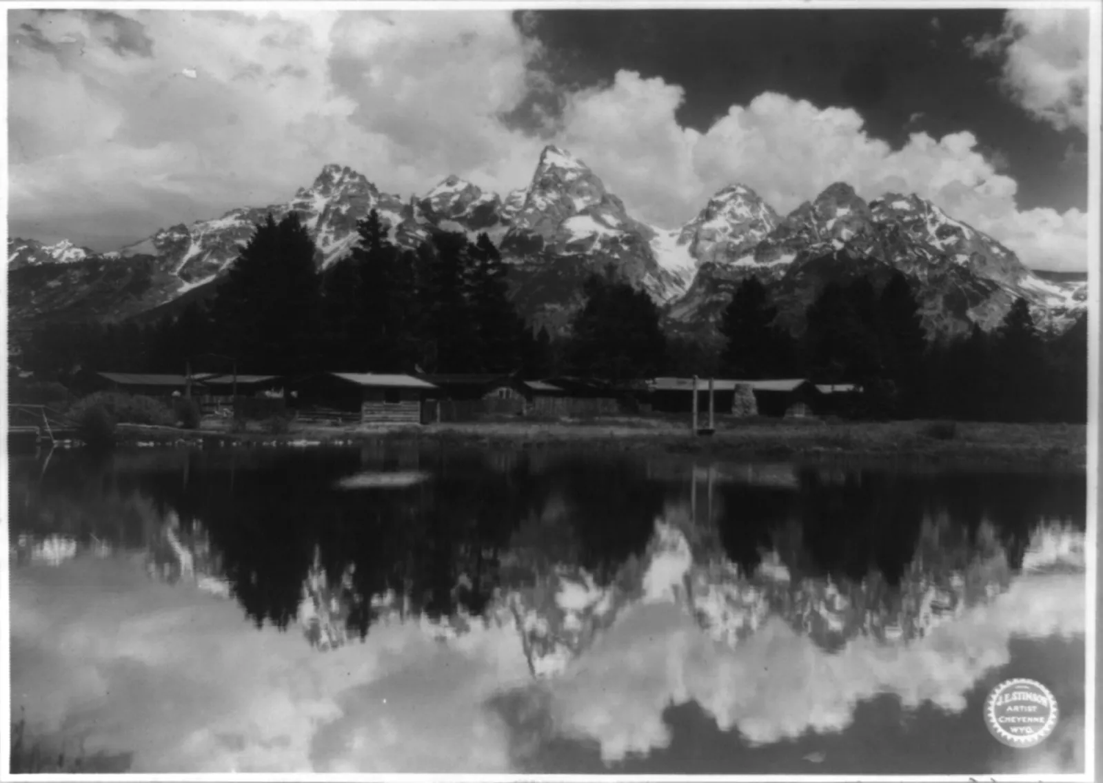The jagged Teton peaks above a line of low ranch buildings, reflected in still water.