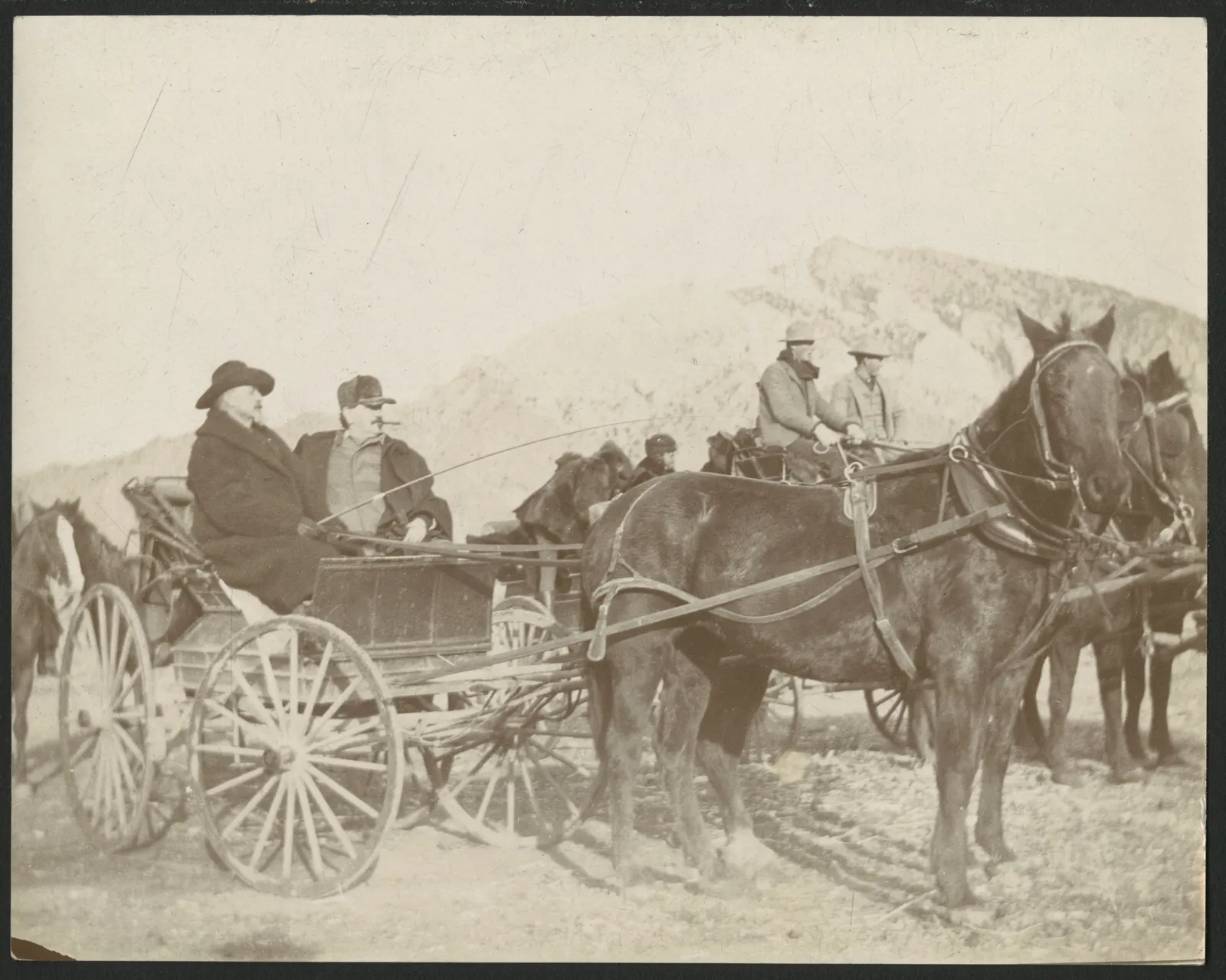 Buffalo Bill Cody and Frank Powell seated in an open carriage behind a team, with a wagon of guests behind them and a mountain beyond.