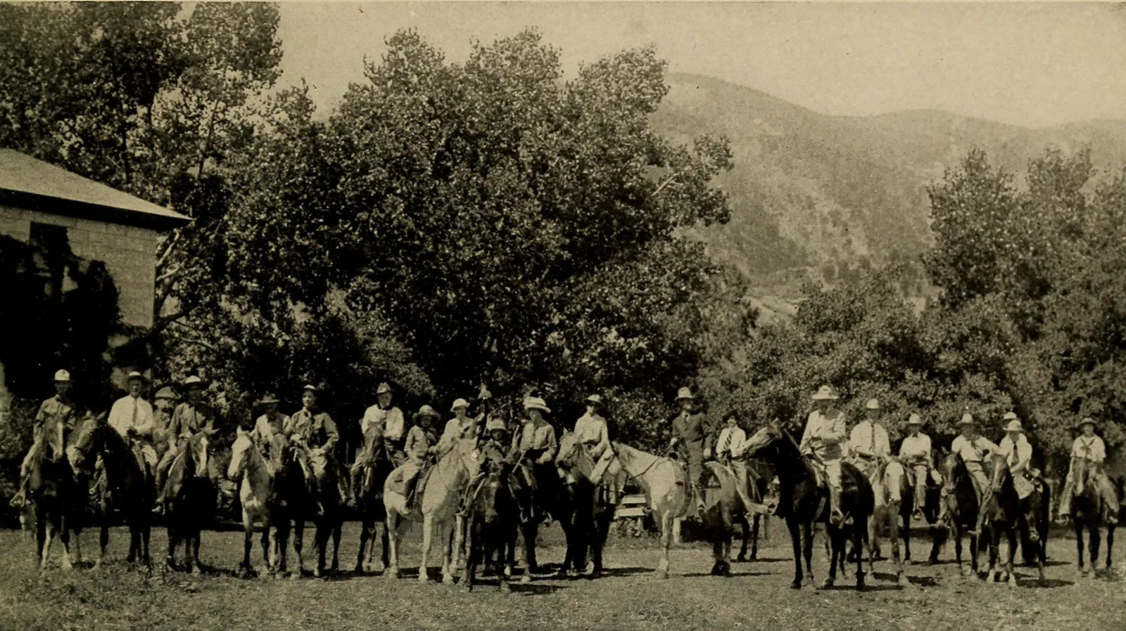 About thirty riders in hats lined up on horseback in front of trees and a ranch building, with mountains behind.