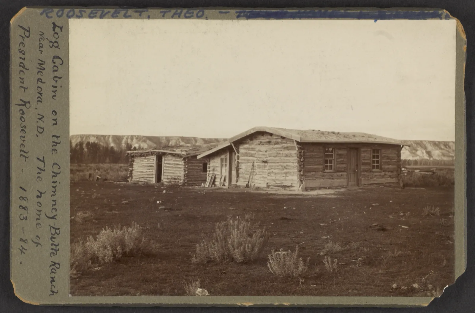 A low log cabin with a shingled roof on open ground near Medora, with a second log building behind it.