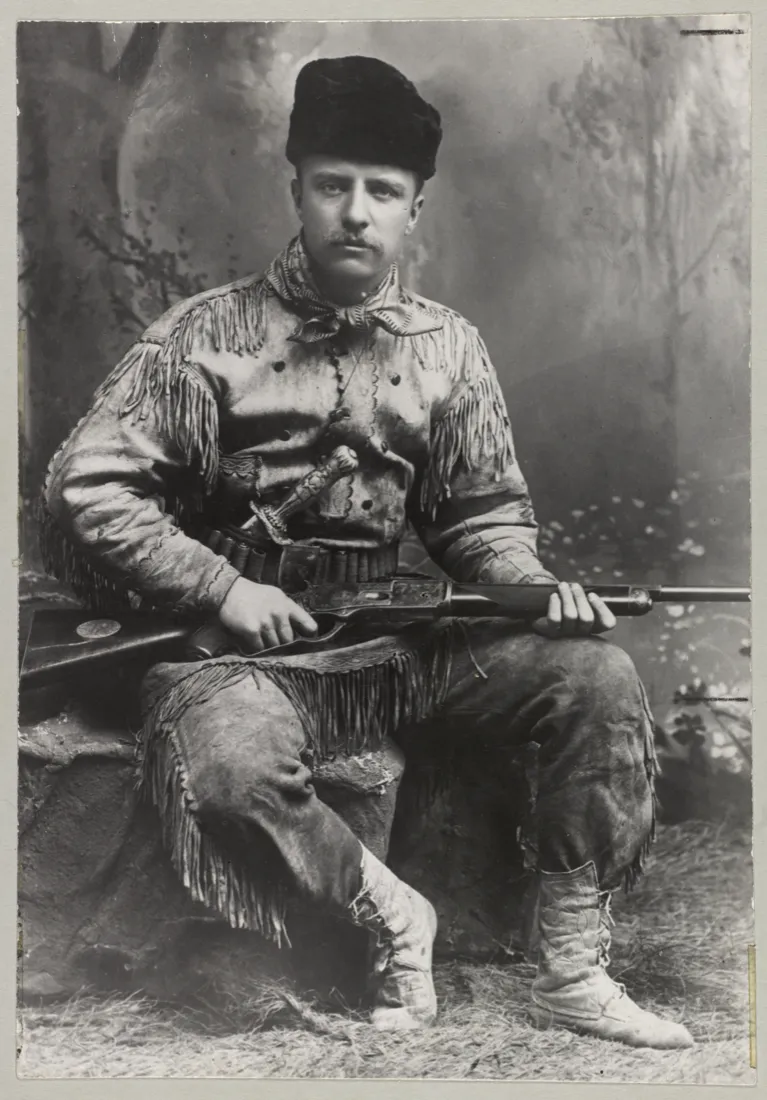 Theodore Roosevelt in a fringed buckskin hunting suit, holding a rifle, in a studio portrait from 1885.