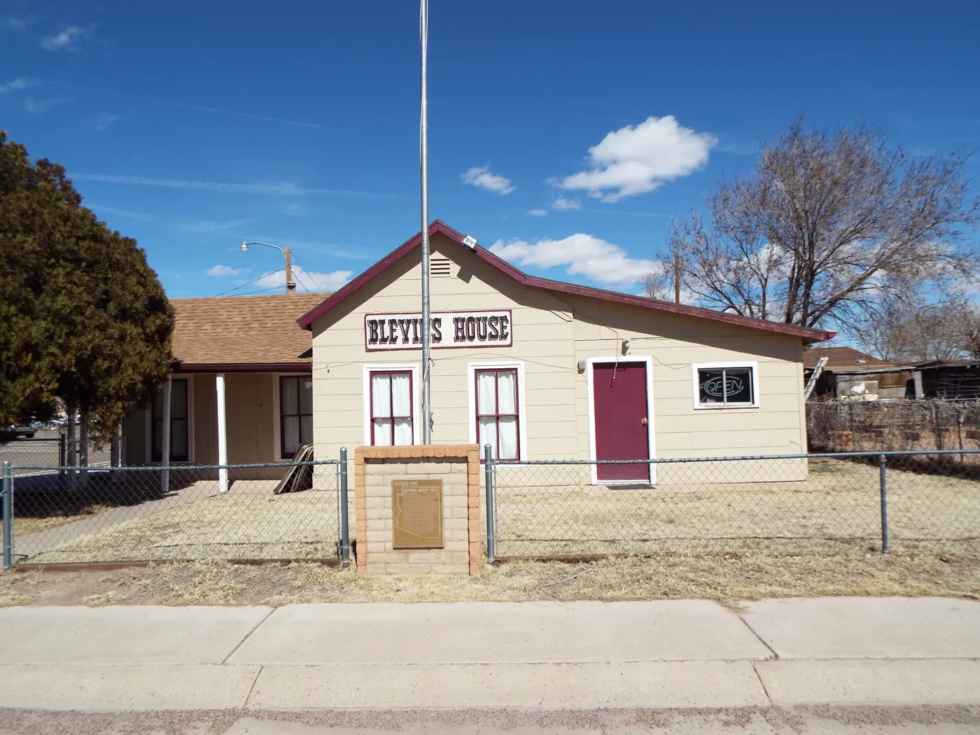 A small tan house with a sign reading Blevins House behind a chain-link fence, with a brick marker post in front.