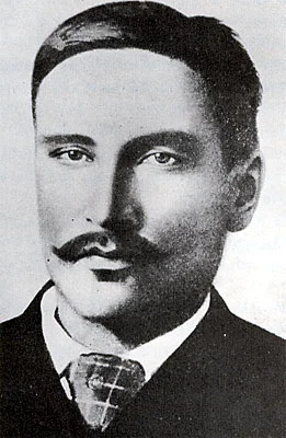 Head-and-shoulders portrait of a young man with a dark moustache, a dark coat and a checked tie.
