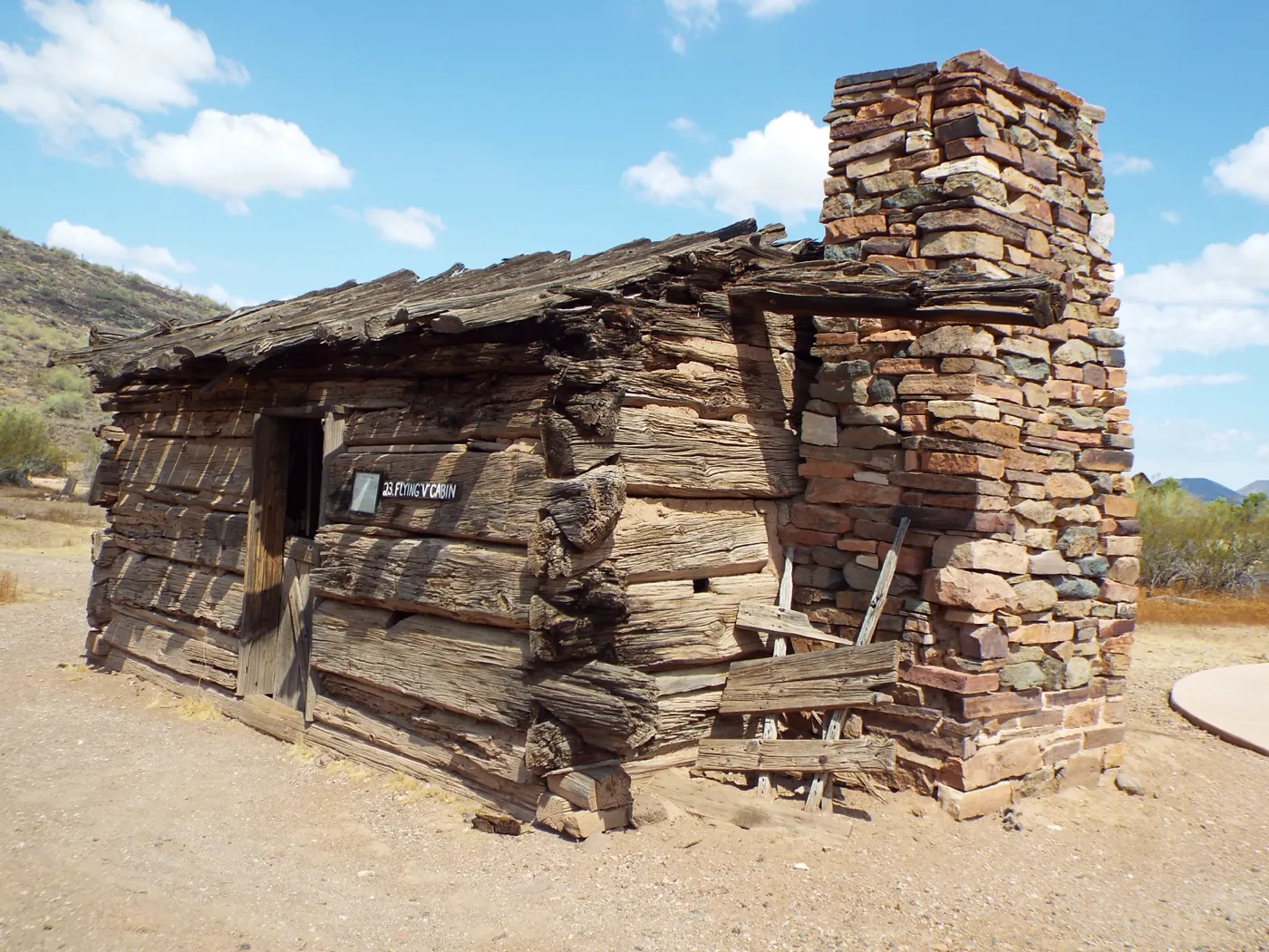 A weathered log cabin with a tall stone chimney on bare desert ground under a blue sky.