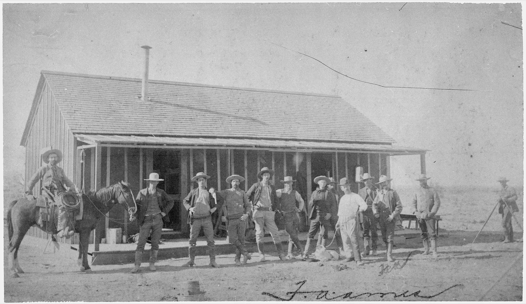 About a dozen cowboys in hats standing in front of a long wooden building with a porch, one man on horseback at left.
