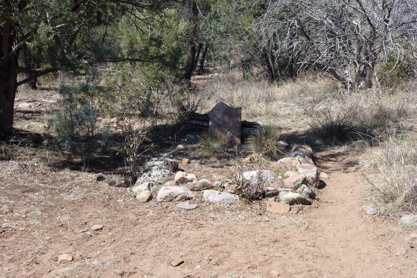 A low ring of stones around a single flat headstone under juniper trees, beside a dirt path.