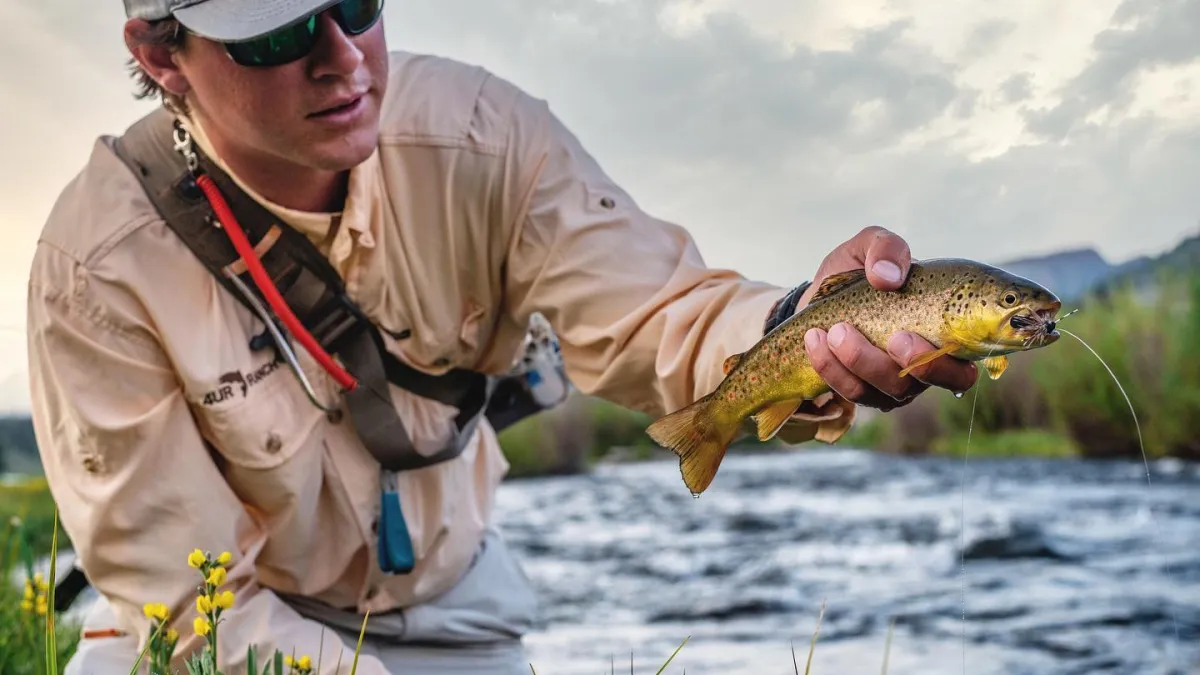 A fishing guide kneeling at the river's edge holding up a brown trout.