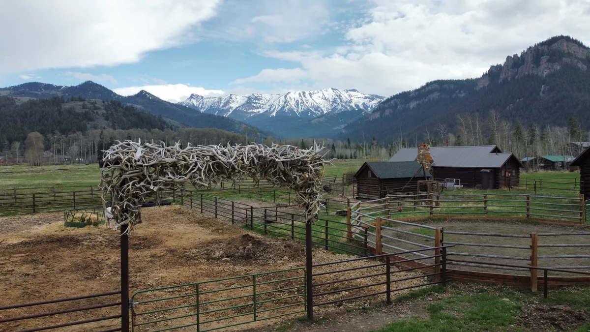 The antler gate and wooden corrals in front of log cabins and snowy peaks.