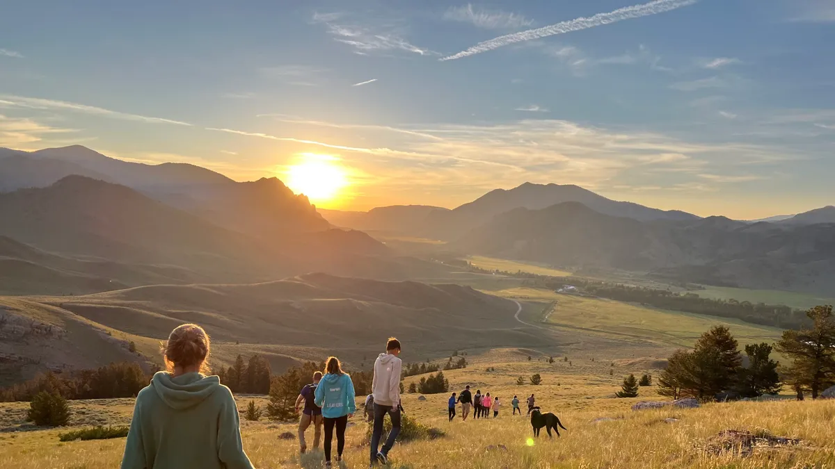 Guests and a dog walking through golden grass toward the sun setting over the mountains.