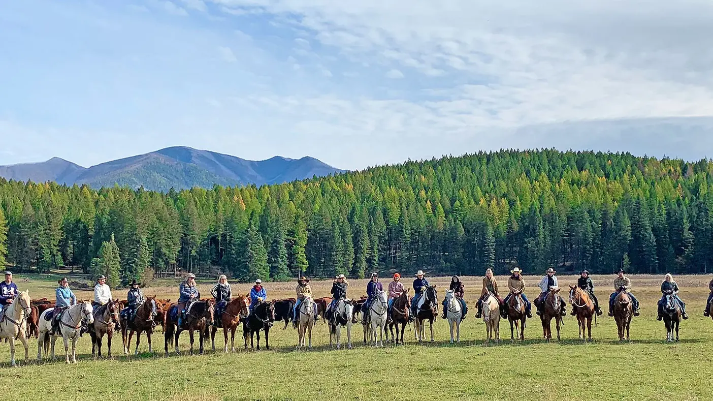 A long line of riders on horseback across a meadow below forested mountains.
