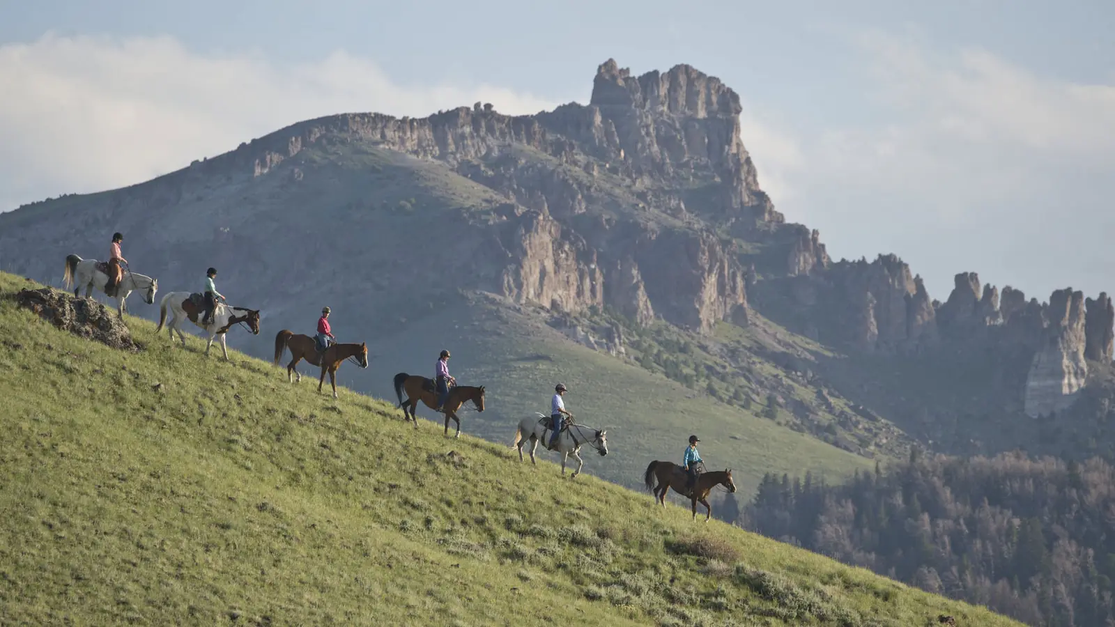 A line of riders crossing a steep green slope below rocky cliffs.