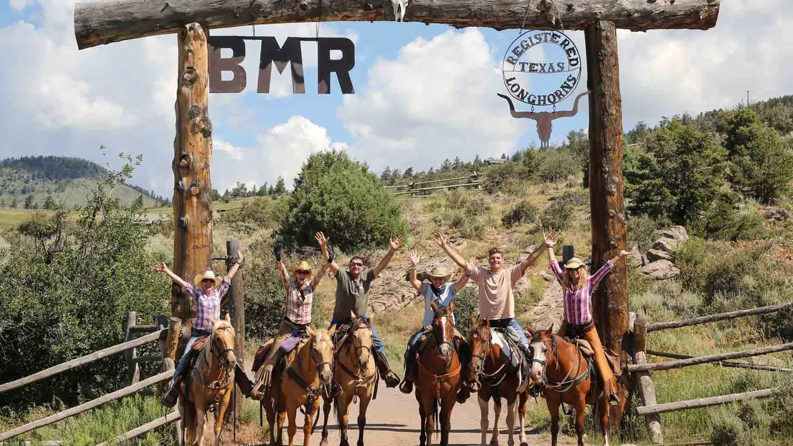 Guests on horseback cheering under the ranch's log entrance sign.