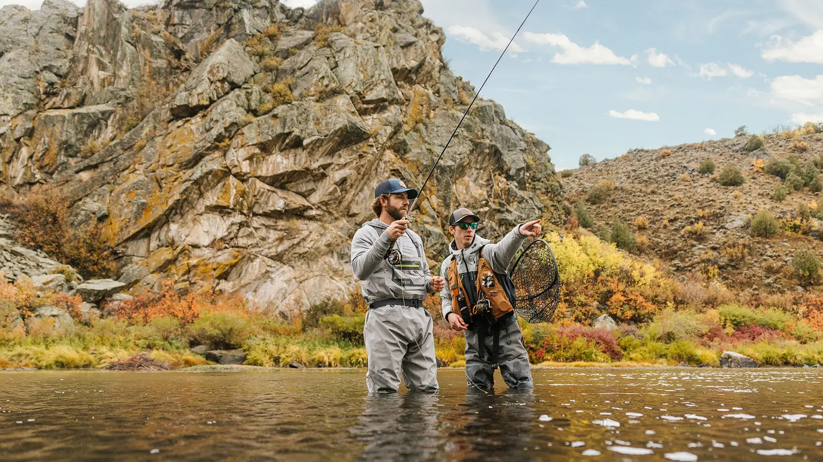 A fly-fishing guide and a guest standing in a river below a rocky cliff.