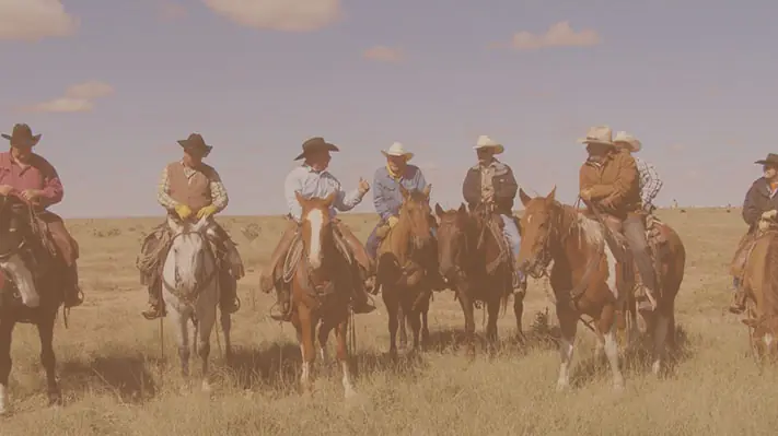 A row of riders on horseback lined up across open New Mexico grassland.