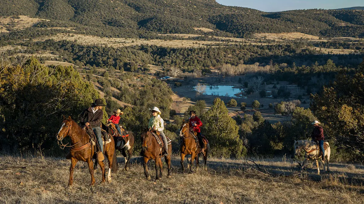 Riders on horseback crossing a slope above a pond in a juniper-covered valley.