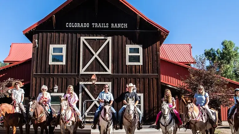 Guests on horseback lined up in front of the ranch's red and brown barn.