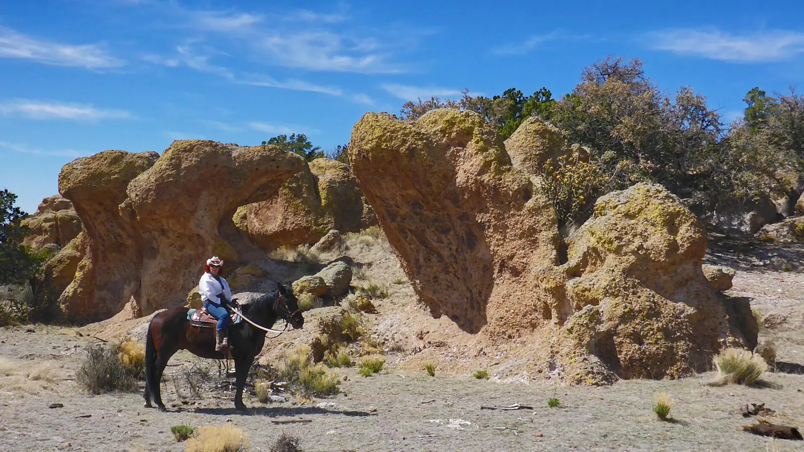 A rider on a black horse beside rounded sandstone boulders under a blue sky.