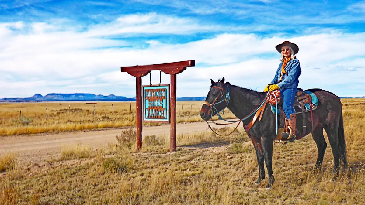 A rider on a dark horse beside the Concho Hills Ranch sign on a gravel road.