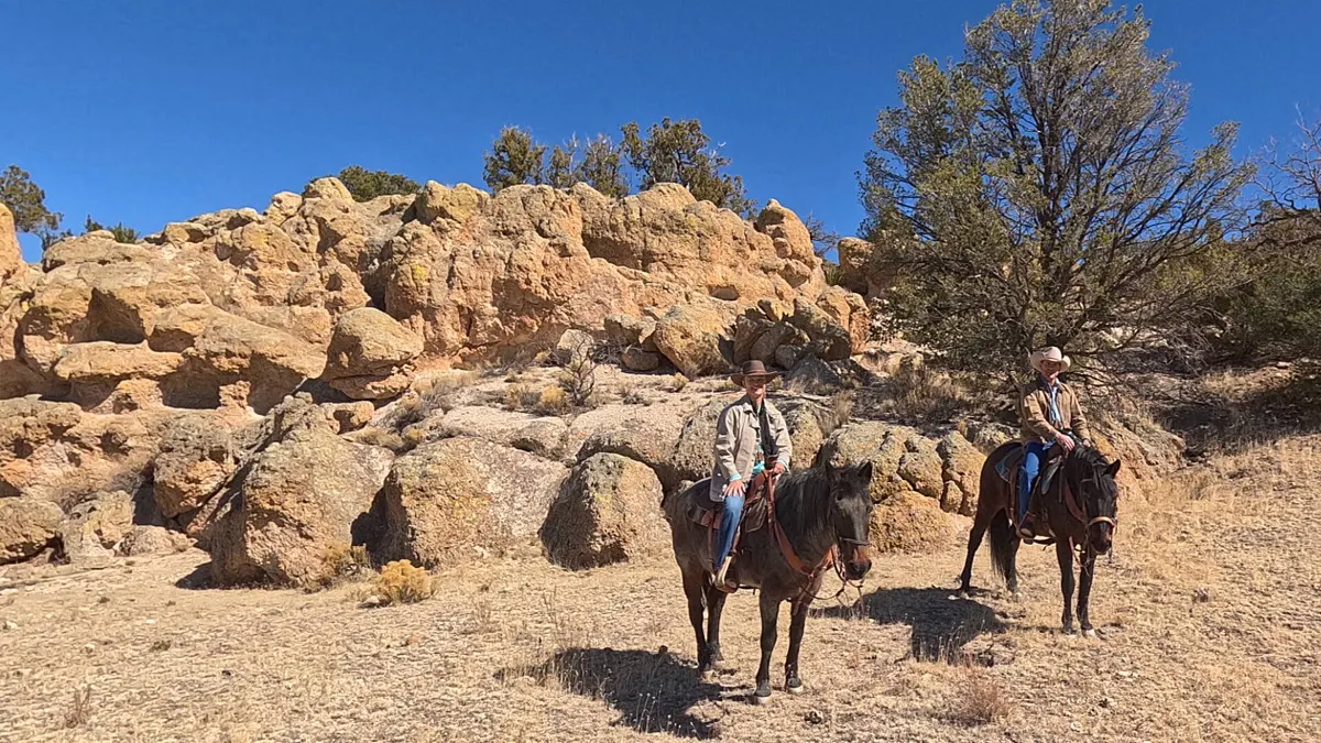 Two riders on horseback among weathered sandstone boulders and juniper trees.