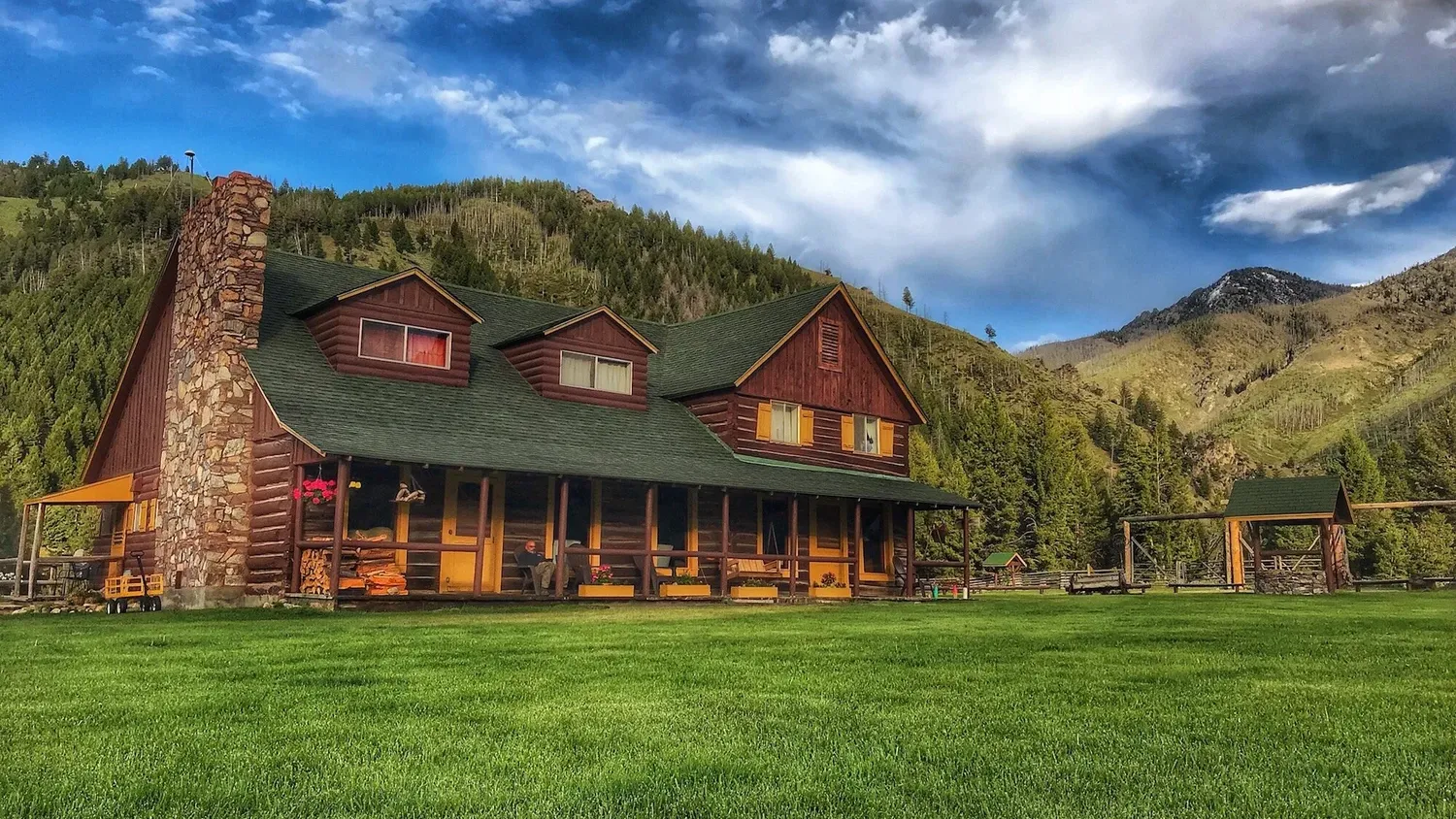 The log lodge with a stone chimney on a green lawn below forested mountains.