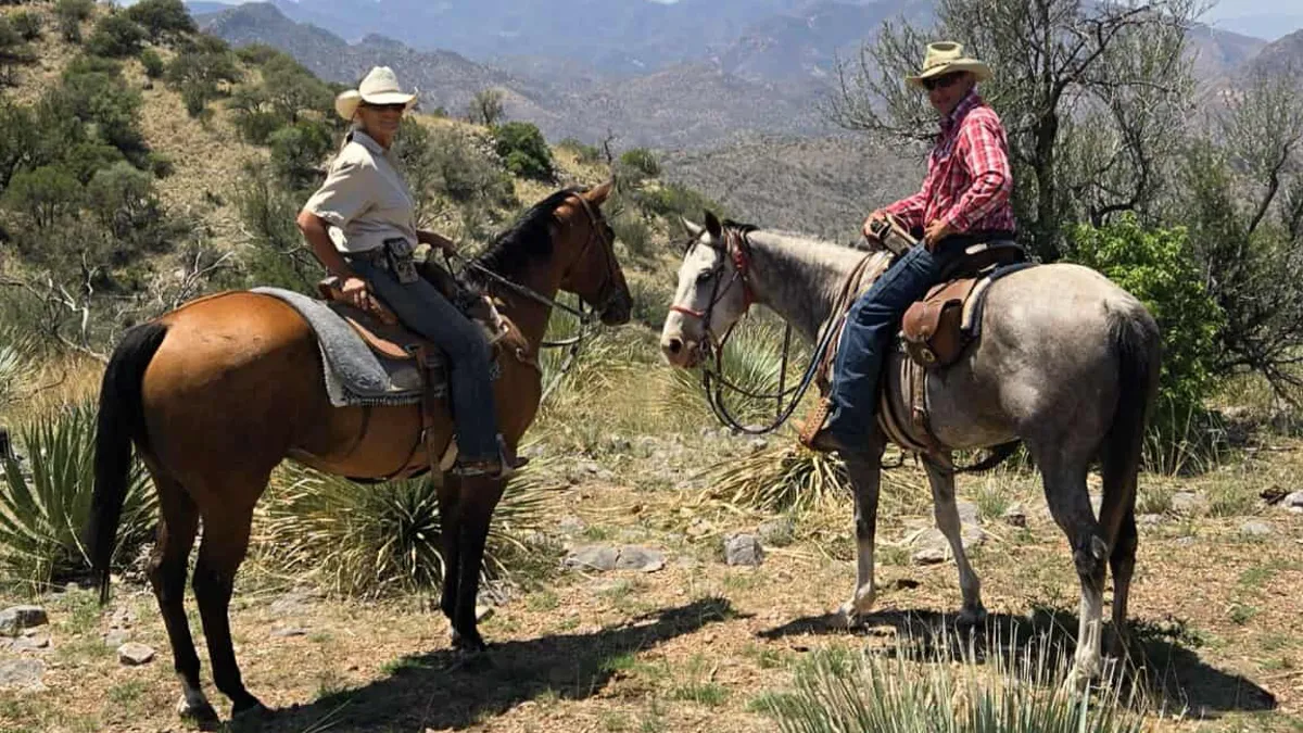 Two riders in straw hats on a bay and a gray horse among yucca, desert mountains behind.