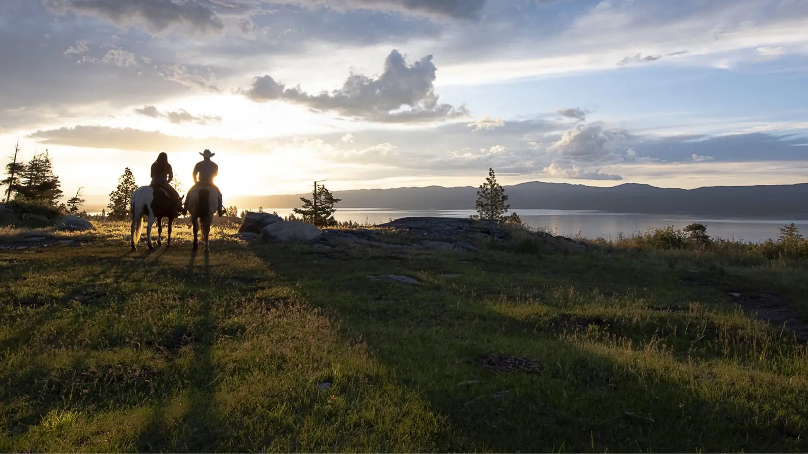 Two riders on horseback on a ridge above Flathead Lake at sunset.