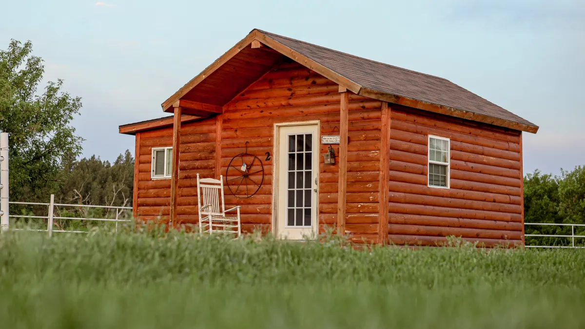 A small log cabin with a rocking chair on the porch, behind a green meadow.