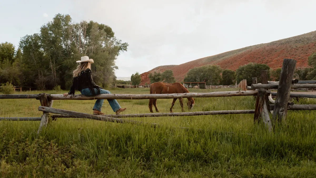 A woman in a cowboy hat sitting on a rail fence while a horse grazes below red hills.