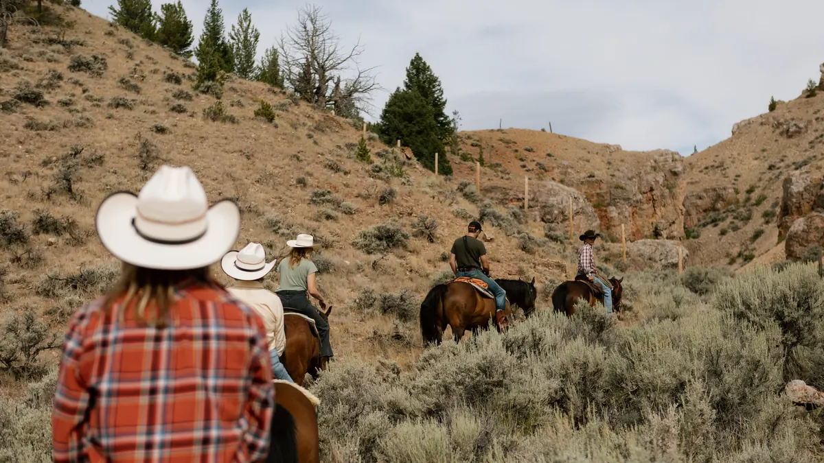 Riders in cowboy hats following a trail through sagebrush toward a rocky draw.