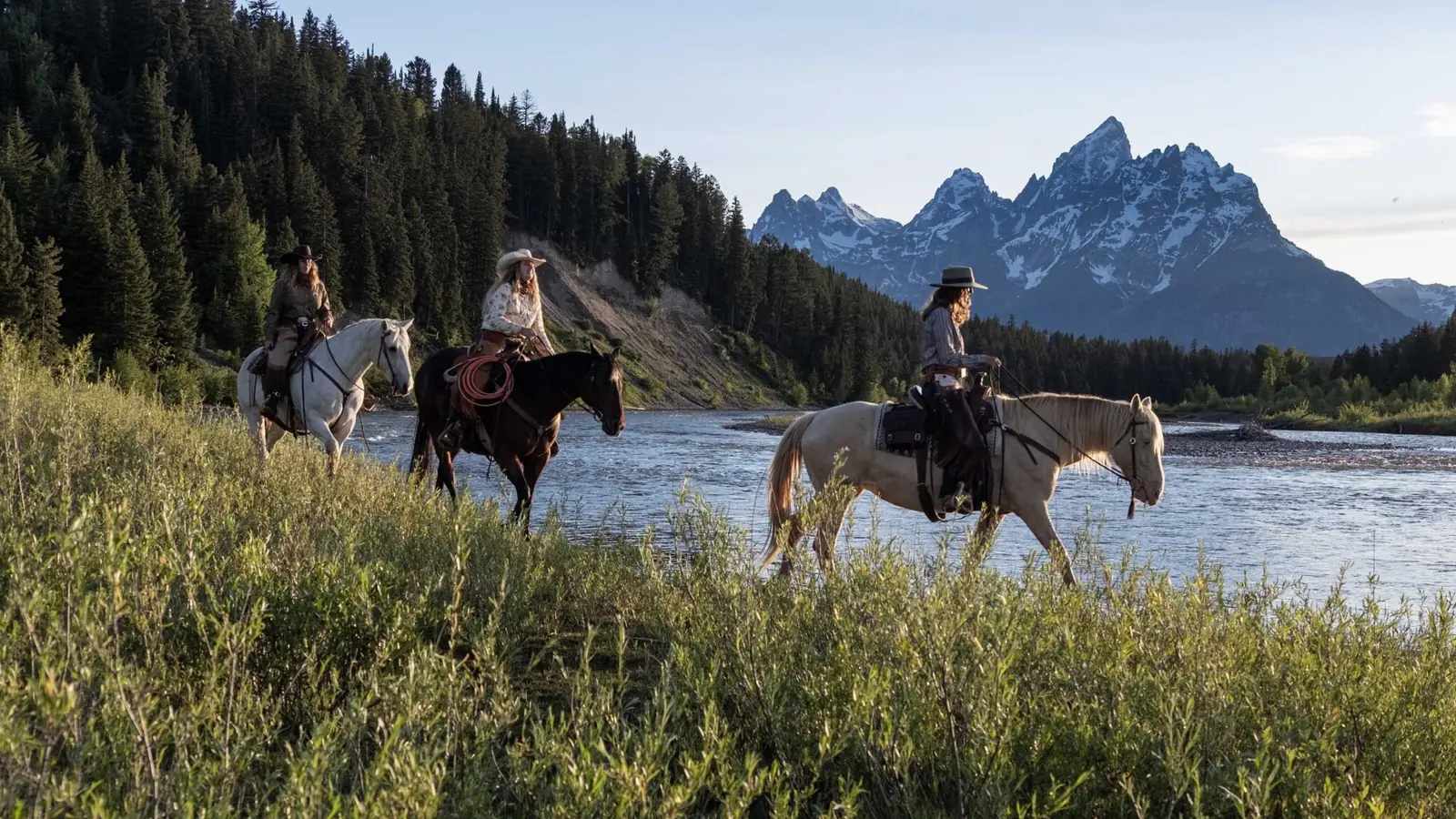 Three riders on horseback beside a river with the Grand Teton behind them.