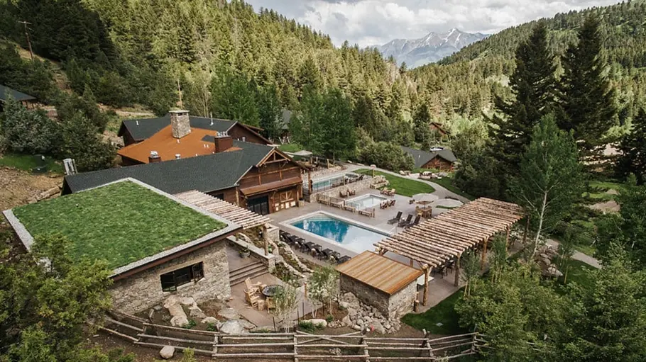 An aerial view of a stone and timber lodge with a pool in a forested mountain valley.