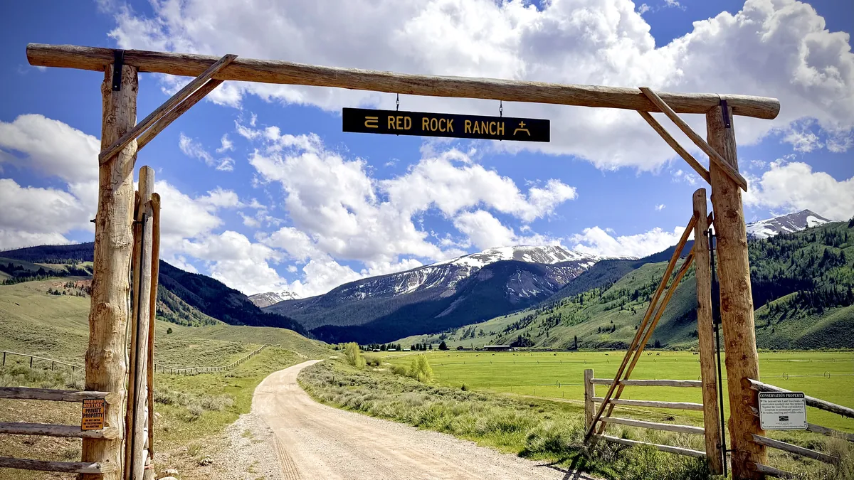 The log entrance gate with the Red Rock Ranch sign, a gravel road leading toward snowy peaks.