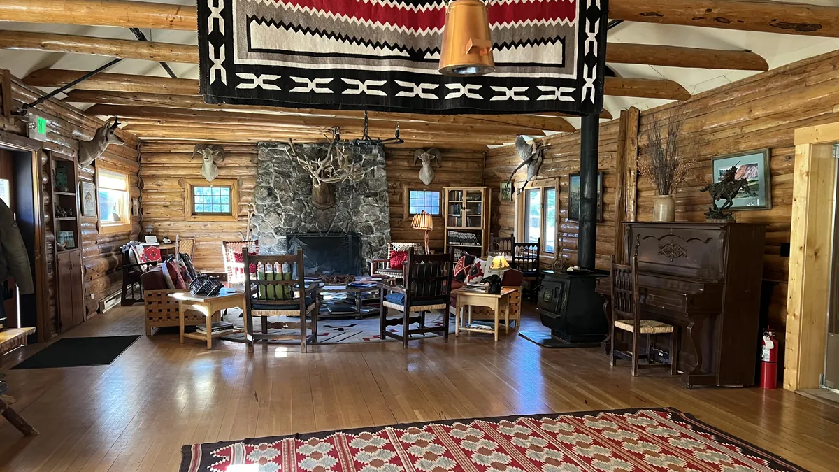 A log-walled lodge room with a stone fireplace, wooden chairs, a piano and patterned rugs.