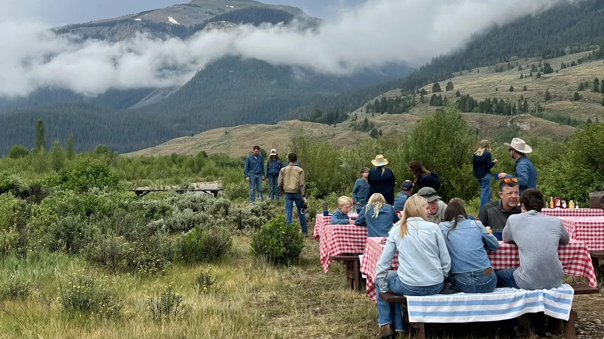 Guests at picnic tables with red checked cloths in a sagebrush meadow, under cloud-wrapped mountains.