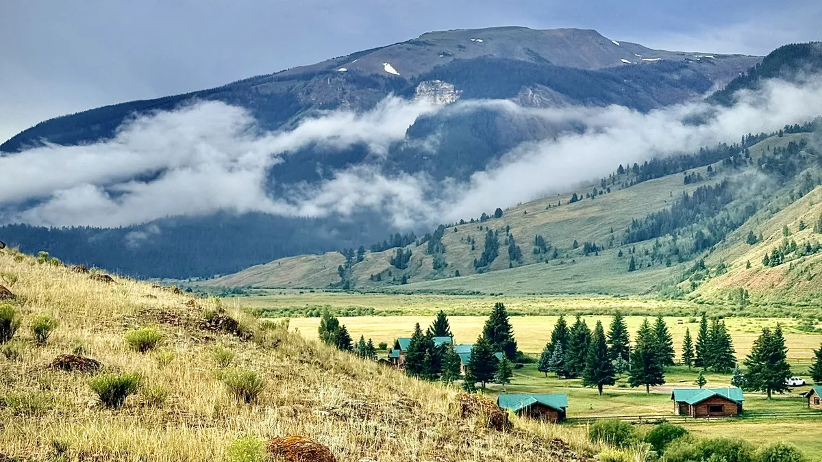 Green-roofed log cabins among pines on the valley floor, with low cloud across the mountain behind.