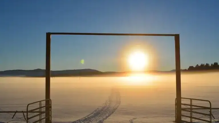 The sun rising through a ranch gate over a misty field.