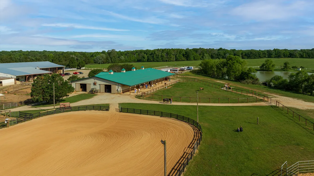 A raked sand arena in front of the green-roofed horse barn, paddocks and a pond.