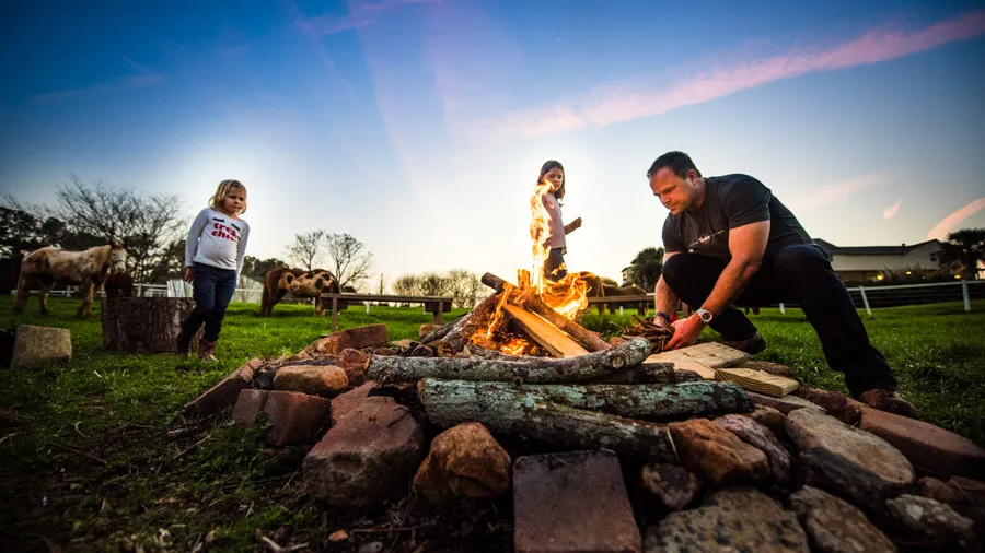 A man building a campfire at dusk while two girls watch, with horses behind.