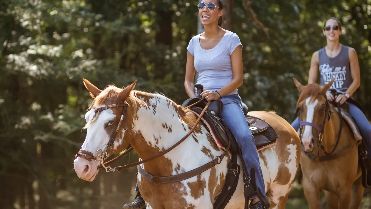 Two smiling women riding a paint and a sorrel horse along a wooded trail.
