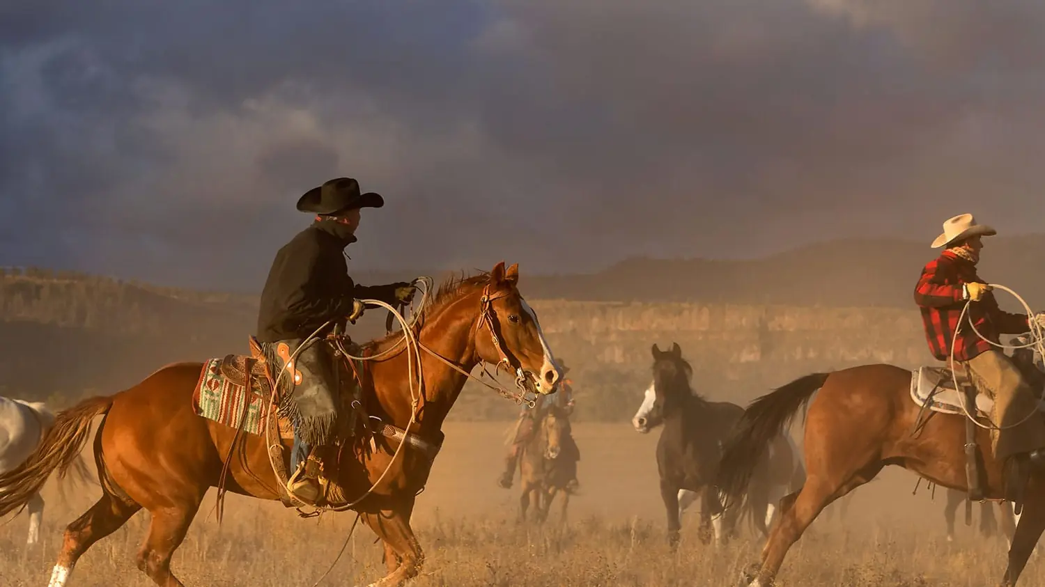 Wranglers loping horses through a dusty meadow under a stormy sky.