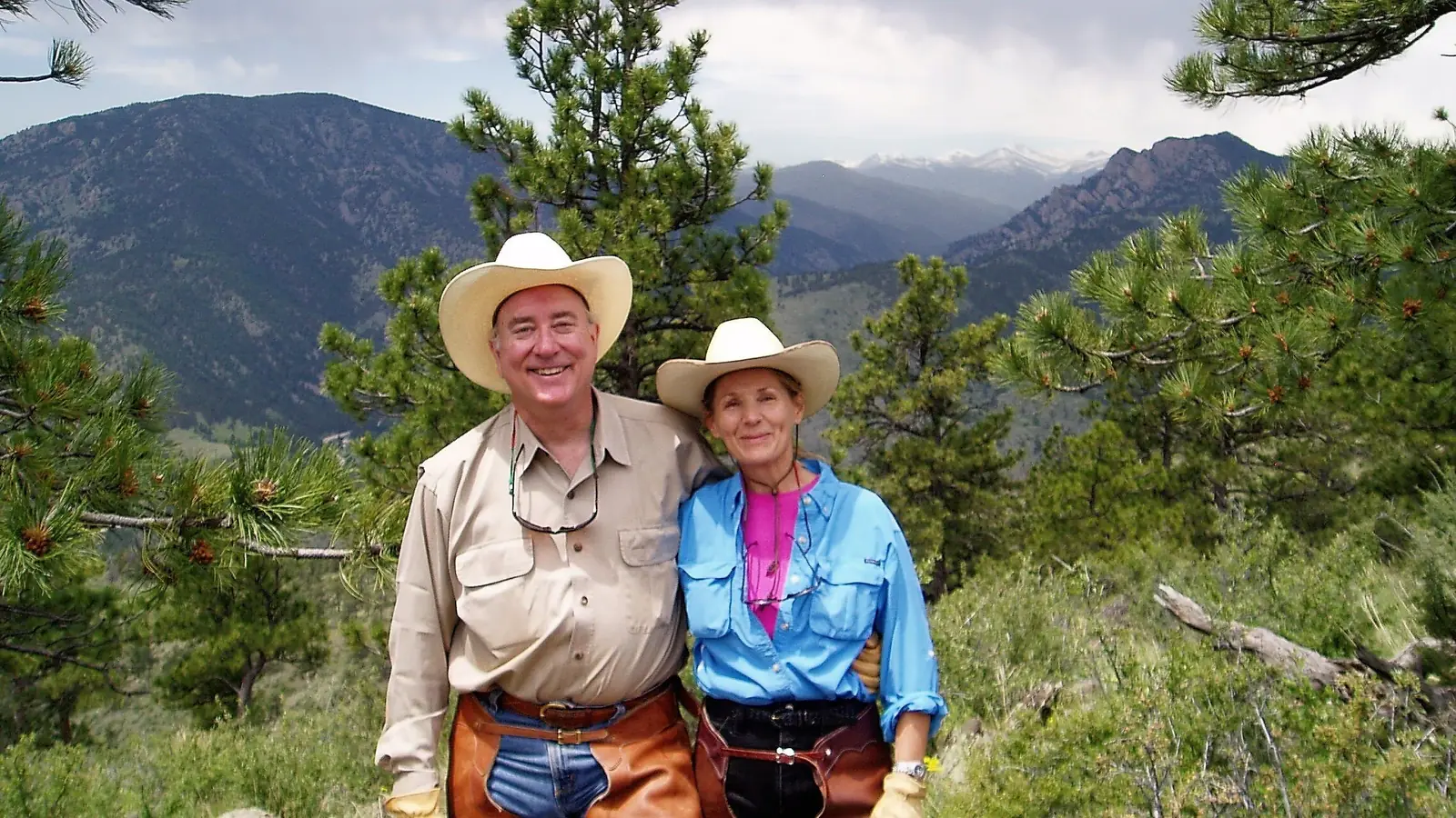 Two ranch hosts in cowboy hats and chaps with pine-covered foothills behind them.