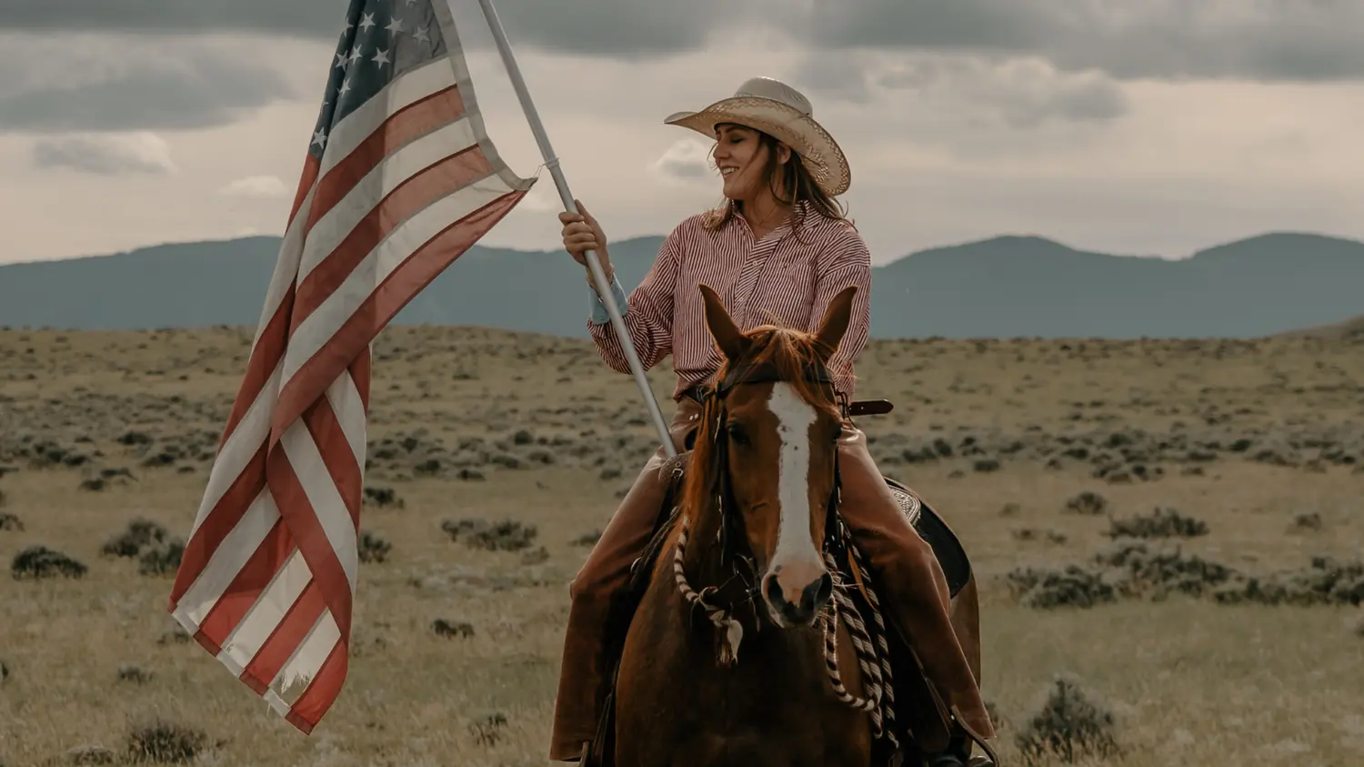 A rider in a straw hat carrying an American flag on horseback across open grassland.