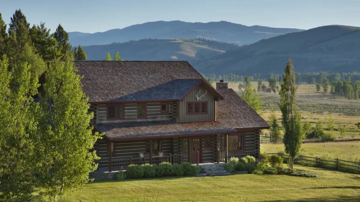 A two-story log house on a green lawn, with rolling hills behind.