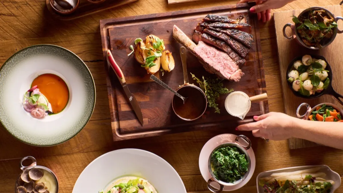 A carved steak on a cutting board, surrounded by sides and sauces on a wooden table.