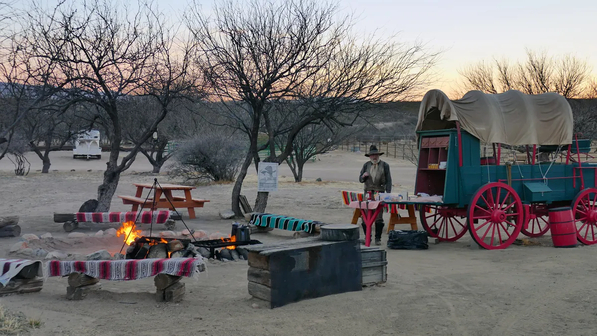 A chuckwagon and campfire breakfast set out under mesquite trees at dawn.