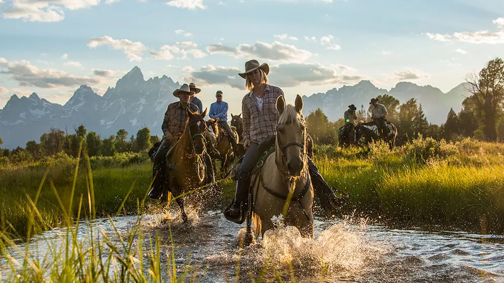 Riders splashing across a creek on horseback with the Teton peaks behind them.